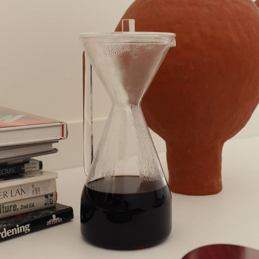 YIELD Pour Over Carafe made of clear borosilicate glass, with a sleek, modern design and a lid for freshness. The carafe features a handle and spout, and is shown with freshly made coffee inside.