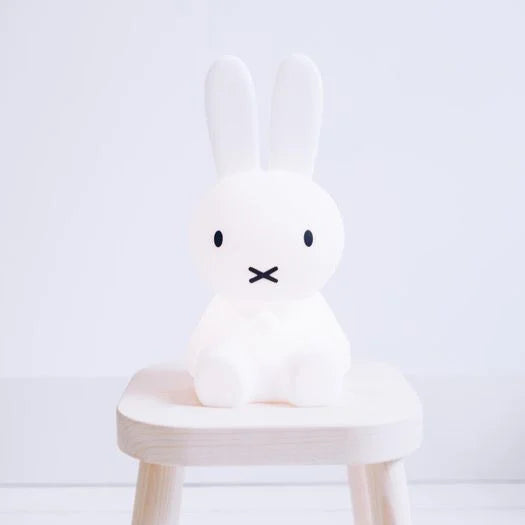 A softly glowing Miffy First Light Lamp sits on a small wooden stool in a minimalist, white room.