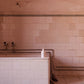 A white bottle of minois bubble bath sits on the rim of a pink tiled square bath that has a purple towel draped over the side of it. Above the bath are two taps and some silver piping against the same pink tiling with a black checkered pattern at the top. The wall above the tiling is also pink with a few cracks in it.