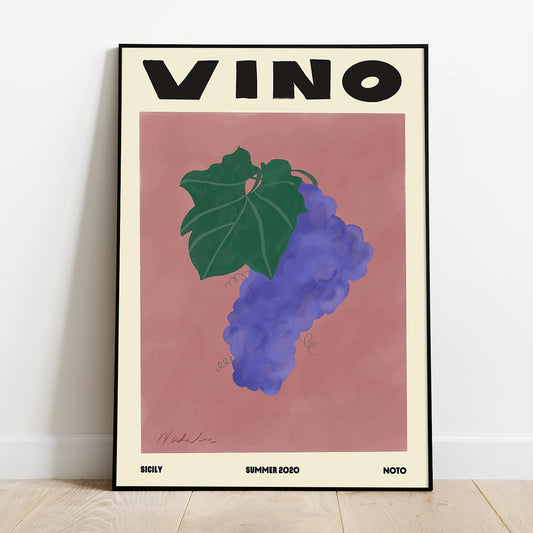 A Natalia Bagniewska A2 Vino print of  purple grapes with dark green leaves on a peach pink background. It says “Vino” which is an informal word for wine. Beneath the grapes it says Sicily on the left hand side in black, Summer 2020 in the middle and Noto on the right hand side. The print is framed and is kept against a white wall on a wooden floor. 