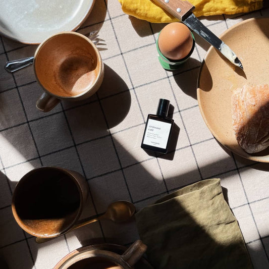 An image of a bottle of the croissant cafe perfume on a white tablecloth with black stripes across it. On the table there is a white plate with a red trim, 2 brown mugs, a gold spoon, a green cloth, a brown knife with butter on it placed against a brown plate with bread on next to a green egg cup with an egg in it. 