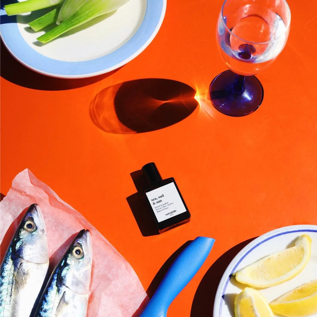 An image of a bottle of perfume, the fragrance being sea, sud & sun, against a red background. There is also a white plate with a blue trim with a green vegetable on it, a clear wine glass with clear liquid inside of it, two fish on top of parchment paper, another white plate with a blue trim that has lemons cut into slices on it and the handle end of a blue knife. 